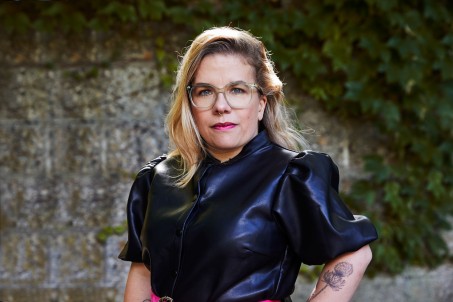 Die Moderatorin Ninia LaGrande at schulterlanges blondes Haar und tr&auml;gt ein Oberteil aus schwarz-blauem Leder mit aufgestelltem Kragen und Puff-&Auml;rmeln. Sie tr&auml;gt pinken Lippenstigt und eine gro&szlig;e graue Brille.
