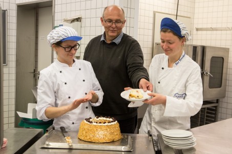 Ein Mann und zwei Frauen in einer Gro&szlig;k&uuml;che. Die beiden Frauen tragen wei&szlig;e Kittel. Eine Frau h&auml;lt einen Teller mit einem St&uuml;ck Kuchen.