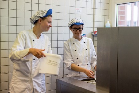 Zwei Frauen in einer Gro&szlig;k&uuml;che. Beide tragen wei&szlig;e Kittel und blau-wei&szlig; gestreifte M&uuml;tzen. Sie stehen hinter einer Bandtransportsp&uuml;lmaschine.