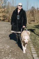 Andrea Eberl geht mit ihrem Blindenhund Enny spazieren. Andrea lacht und tr&auml;gt eine Sonnenbrille. Enny ist ein Golden Retriever und l&auml;uft im Geschirr.