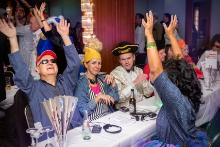 Man sieht eine Gruppe sehbehinderter Jecken, die zusammen an einer Tisch sitzen und jubelnd die H&auml;nde in die H&ouml;he halten.